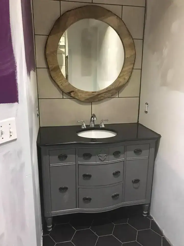 Gray vanity with a dark top and round wood mirror on a tiled wall